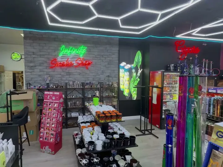 Green and red Infinity Smoke Shop neon sign on a stone wall above a table of coffee mugs