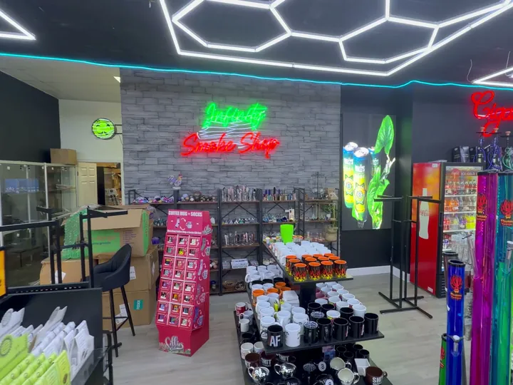 Green and red Infinity Smoke Shop neon sign on a stone wall above shelves of crystals and mugs