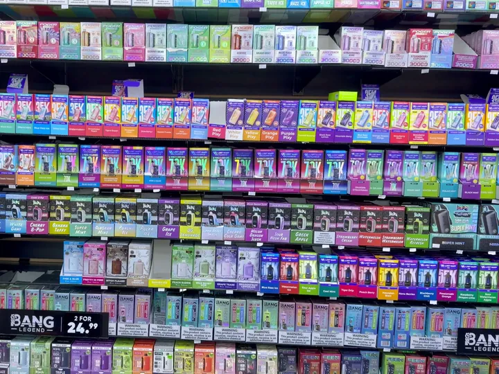 Wall of boxed disposable vapes in rainbow colours on black shelving, cigarette packs at left