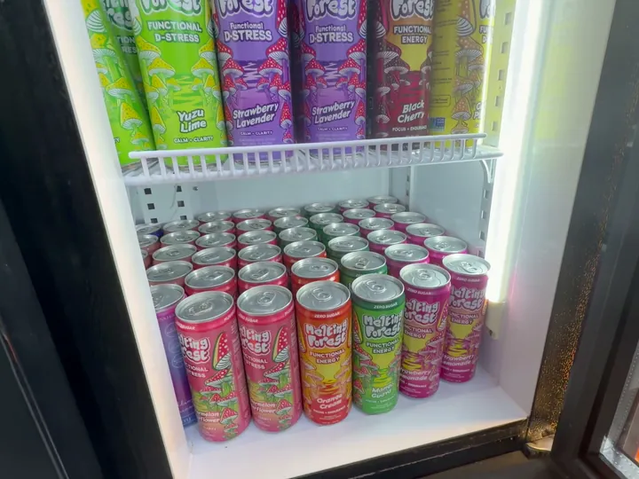Open drink fridge with brightly illustrated cans on a wire shelf and rows of cans below